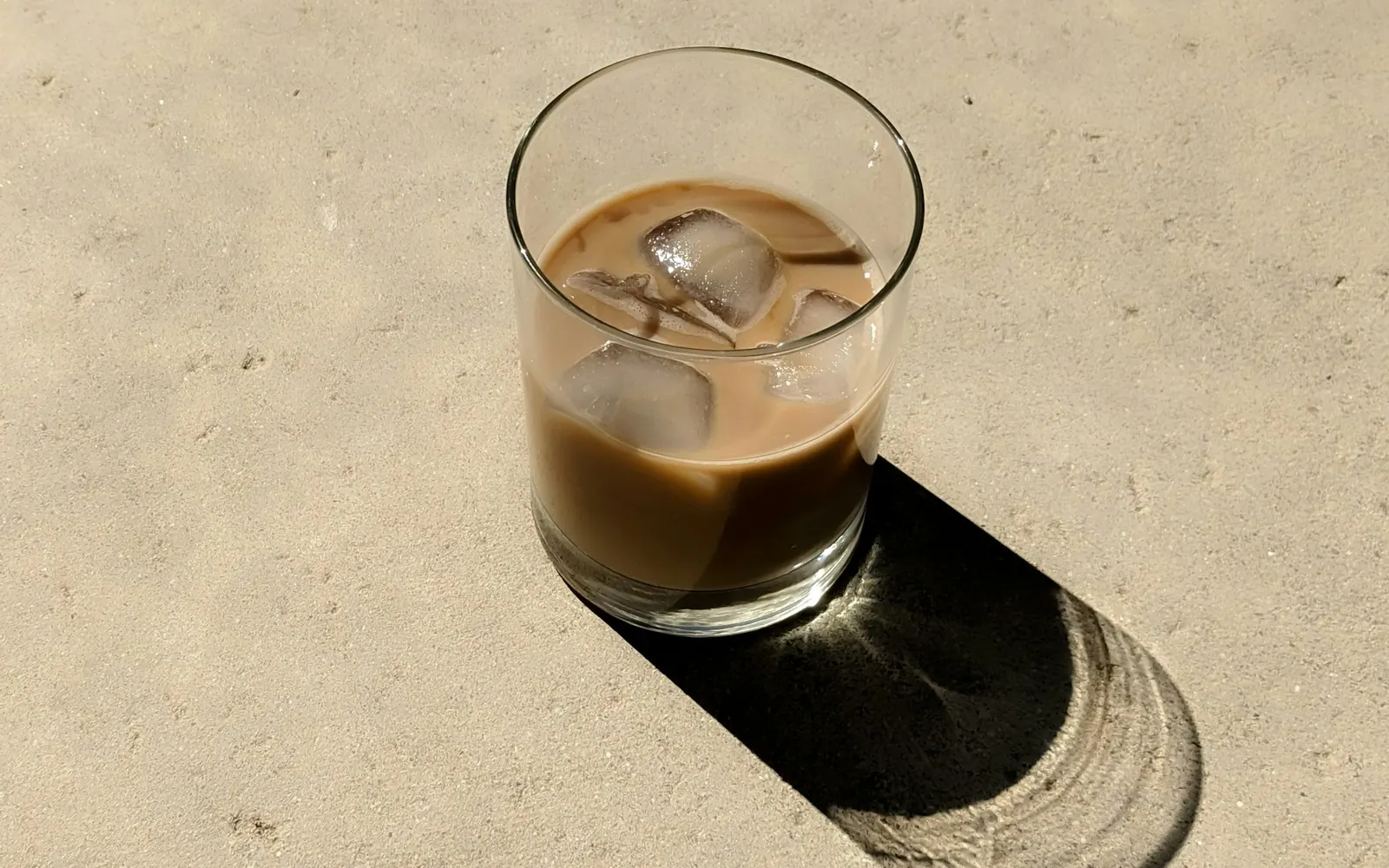 Eiskaffee mit Milch und Eiswürfeln in einem Glas auf hellem Beton, mit langem Schatten