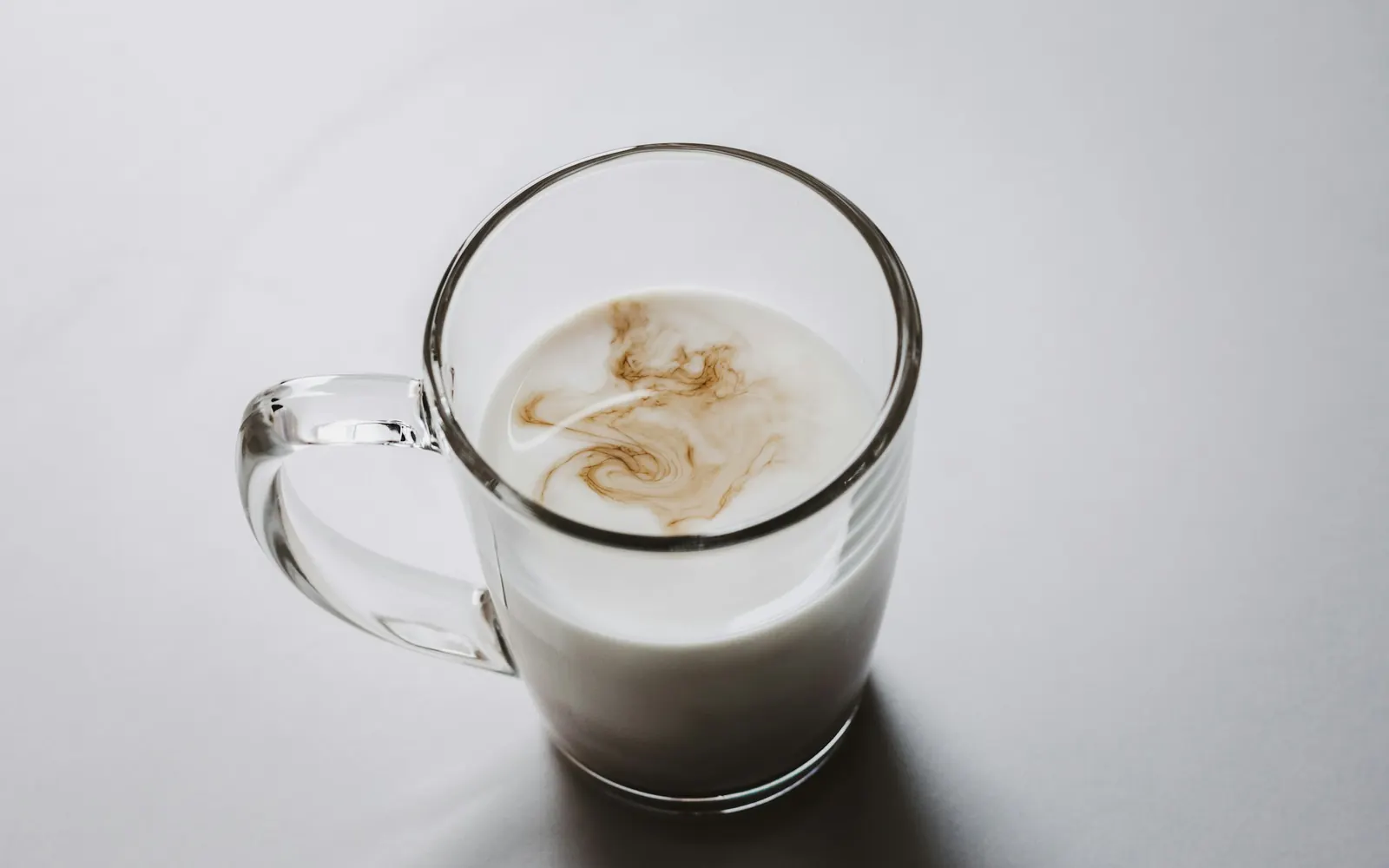 Glasbecher mit Milch, in die sich Kaffee in feinen Schlieren mischt, vor grauem Hintergrund