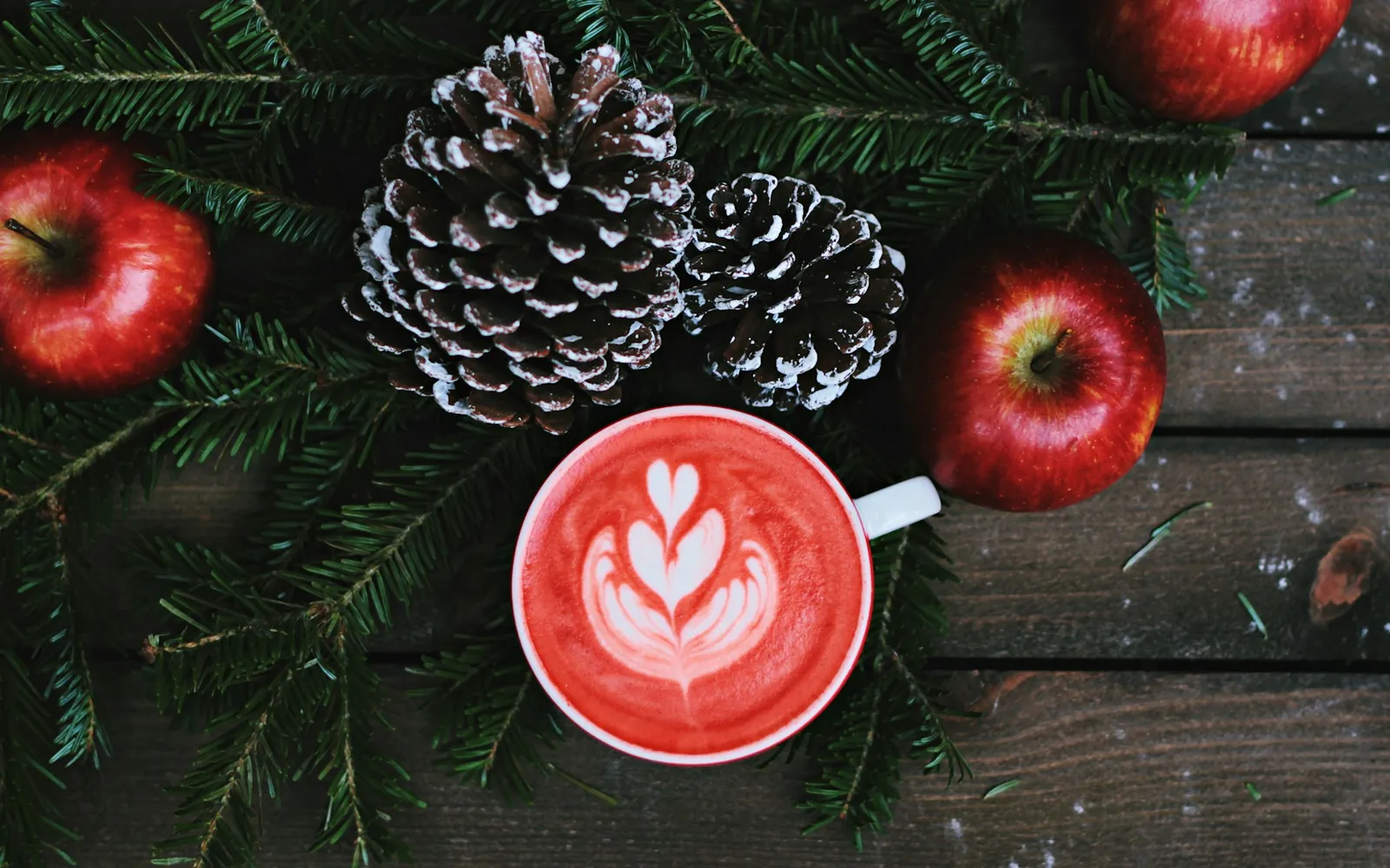 Rote Tasse Latte mit Latte Art zwischen Tannenzweigen, Tannenzapfen und roten Äpfeln